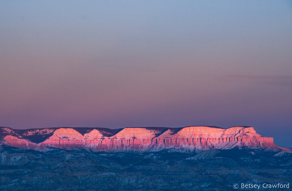 Against a darkening blue and pink sky, a long red rock ridge in southern Utah is lit by the setting sun, shining bright pink over the shadowy land below. Photo by Betsey Crawford.
