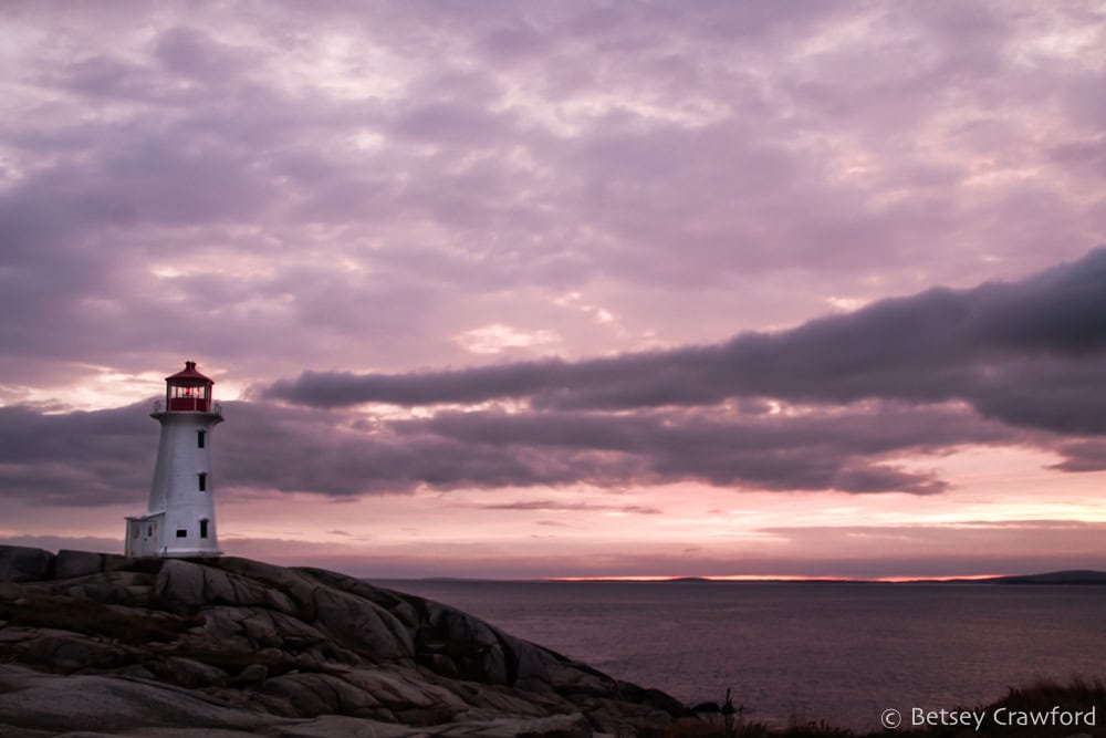 A lighthouse stands on a ridge overlooking the ocean against a backdrop of a sky full of clouds still lit by a recent sunset. Photo by Betsey Crawford.
